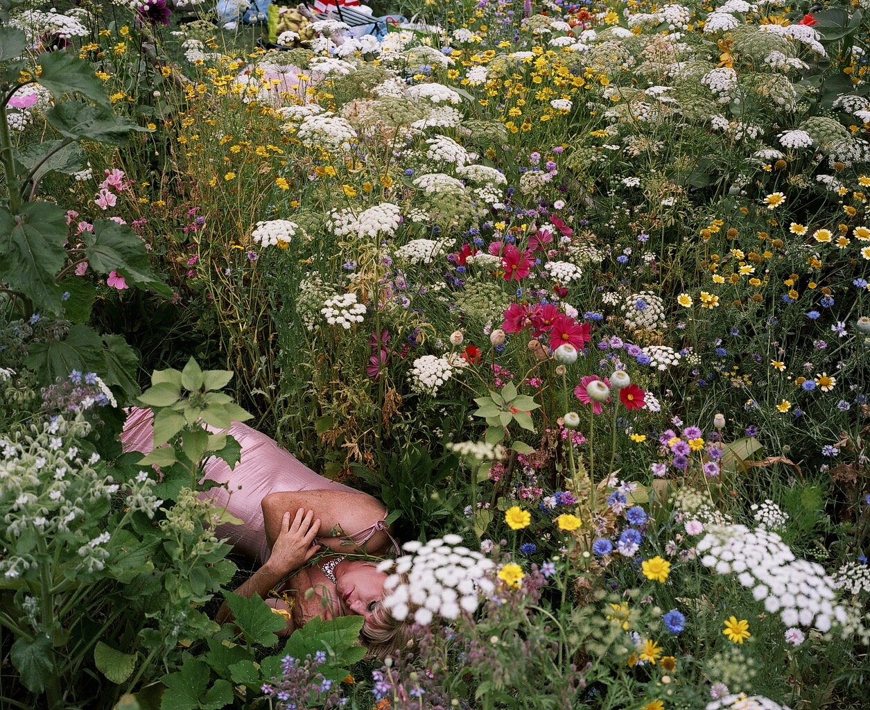 Siân Davey - a woman lying in a pink dress on the ground of a garden with flowers and green plants
