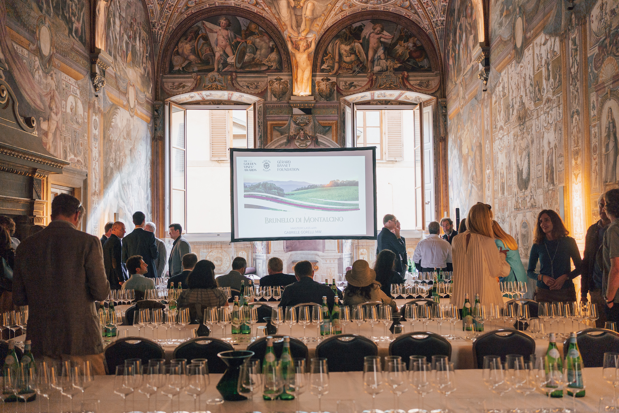 Golden Vines -people in a grand italian room looking at a screen with a set table behind them