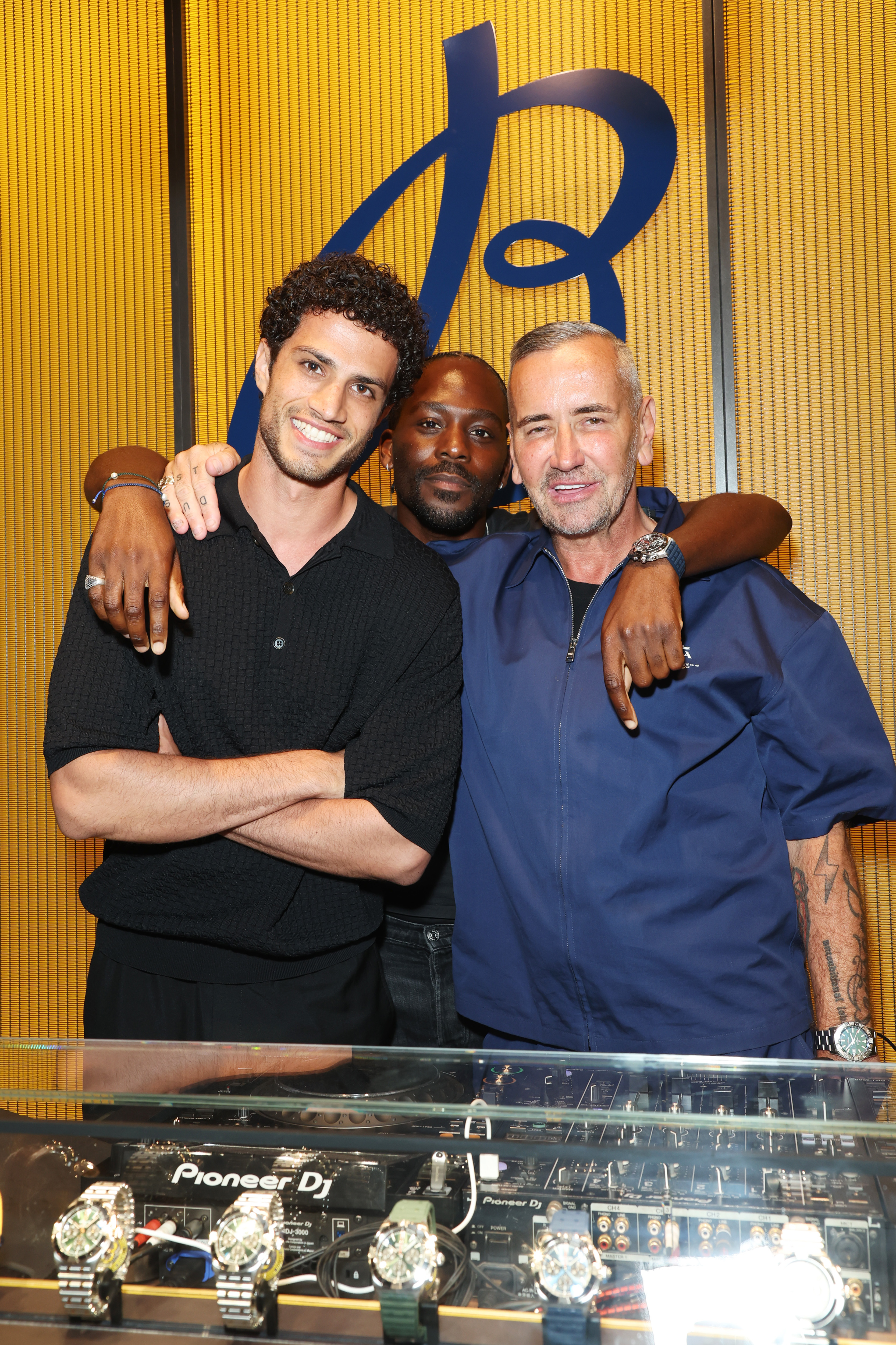 Breitling - three men standing in front of a yellow wall with a big B behind them