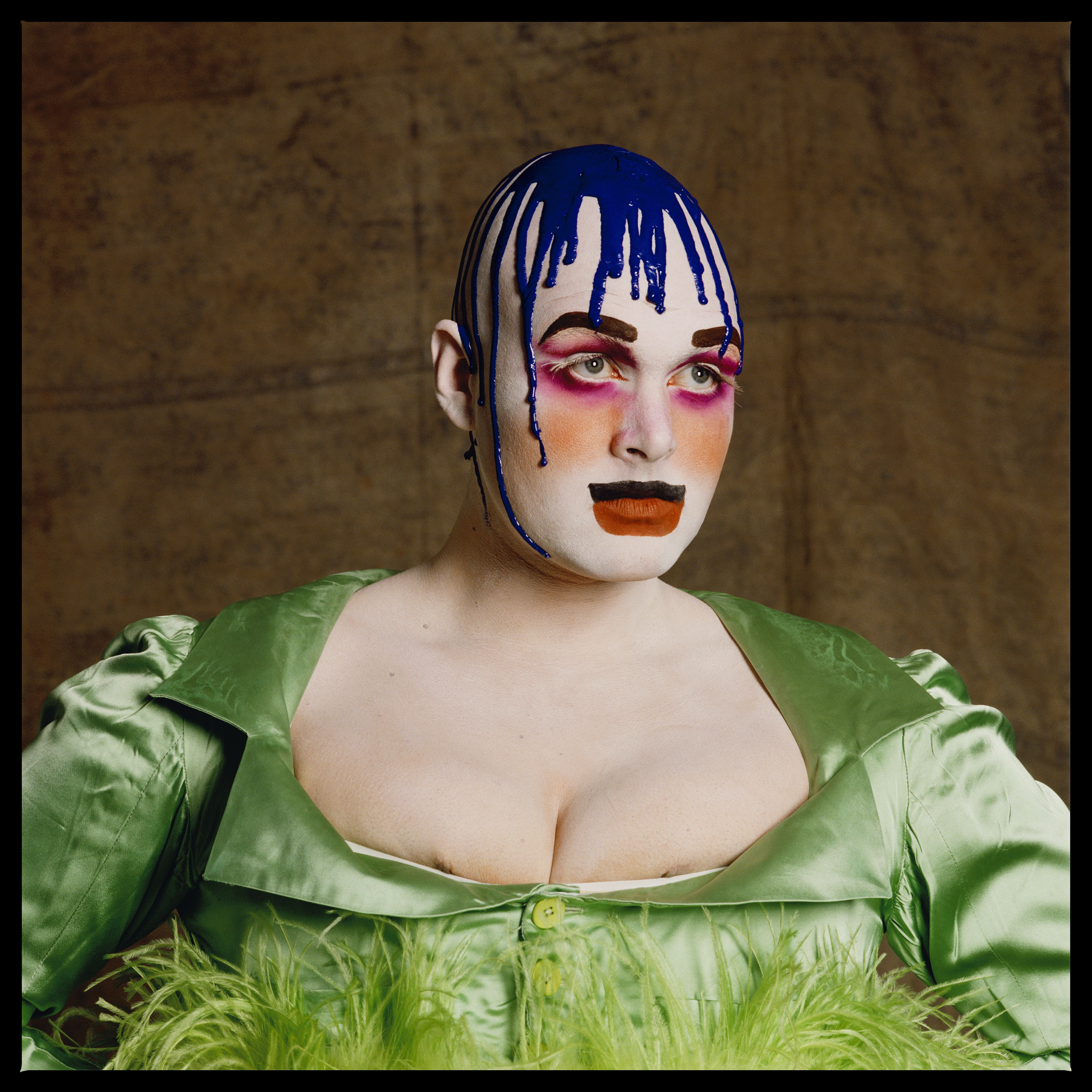 Leigh Bowery Tate Modern - Leigh Bowery in extravagant makeup and fashion. The individual wears a green satin dress with feather accents, with bold makeup featuring bright pink eyeshadow, a blue painted drip effect running down their bald head, and a striking orange mouth. The background is a textured neutral colour, adding to the dramatic and avant-garde look of the image