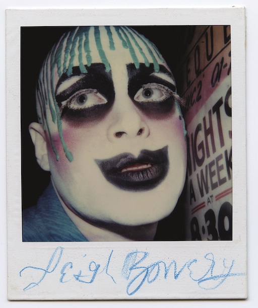 Leigh Bowery Tate Modern - A Polaroid photograph of Leigh Bowery, showing him in dramatic clown-like makeup. His face is painted with white and black makeup, with blue streaks dripping from his bald head. The makeup is exaggerated with bold, dark eye shadow and a large, painted smile. Below the image, Leigh Bowery's signature is written in blue crayon. The background includes part of a sign that reads "NIGHTS A WEEK." The photo captures the avant-garde and theatrical essence of Bowery's persona