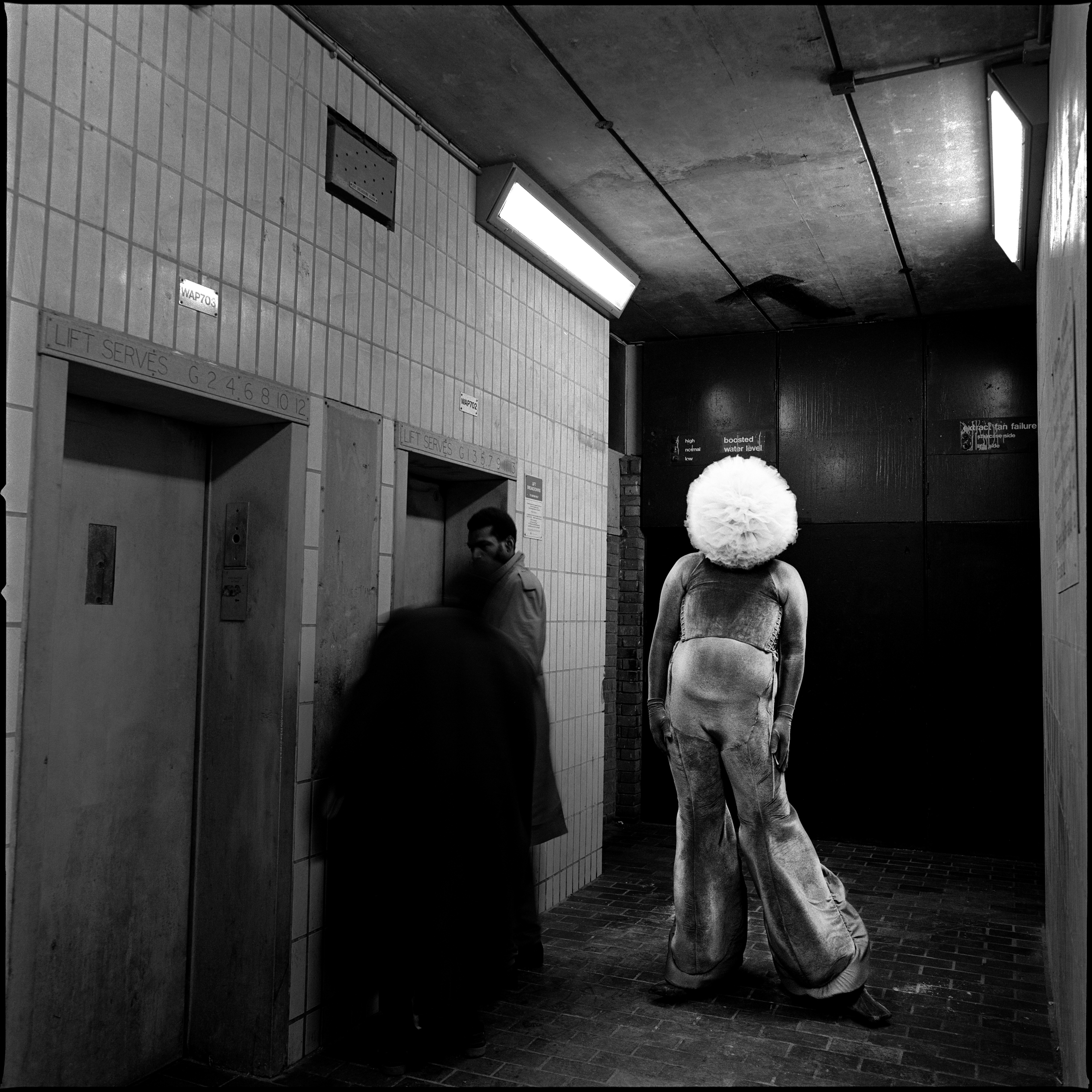 Leigh Bowery Tate Modern - A black-and-white photograph of Leigh Bowery, wearing an oversized white wig and a distressed, dramatic outfit. He stands in a stark, industrial corridor near an elevator, striking a theatrical pose with his back to the camera. Two figures can be seen walking by in the background, adding to the surreal atmosphere of the image. The contrast between Bowery's extravagant appearance and the mundane setting highlights his avant-garde style.