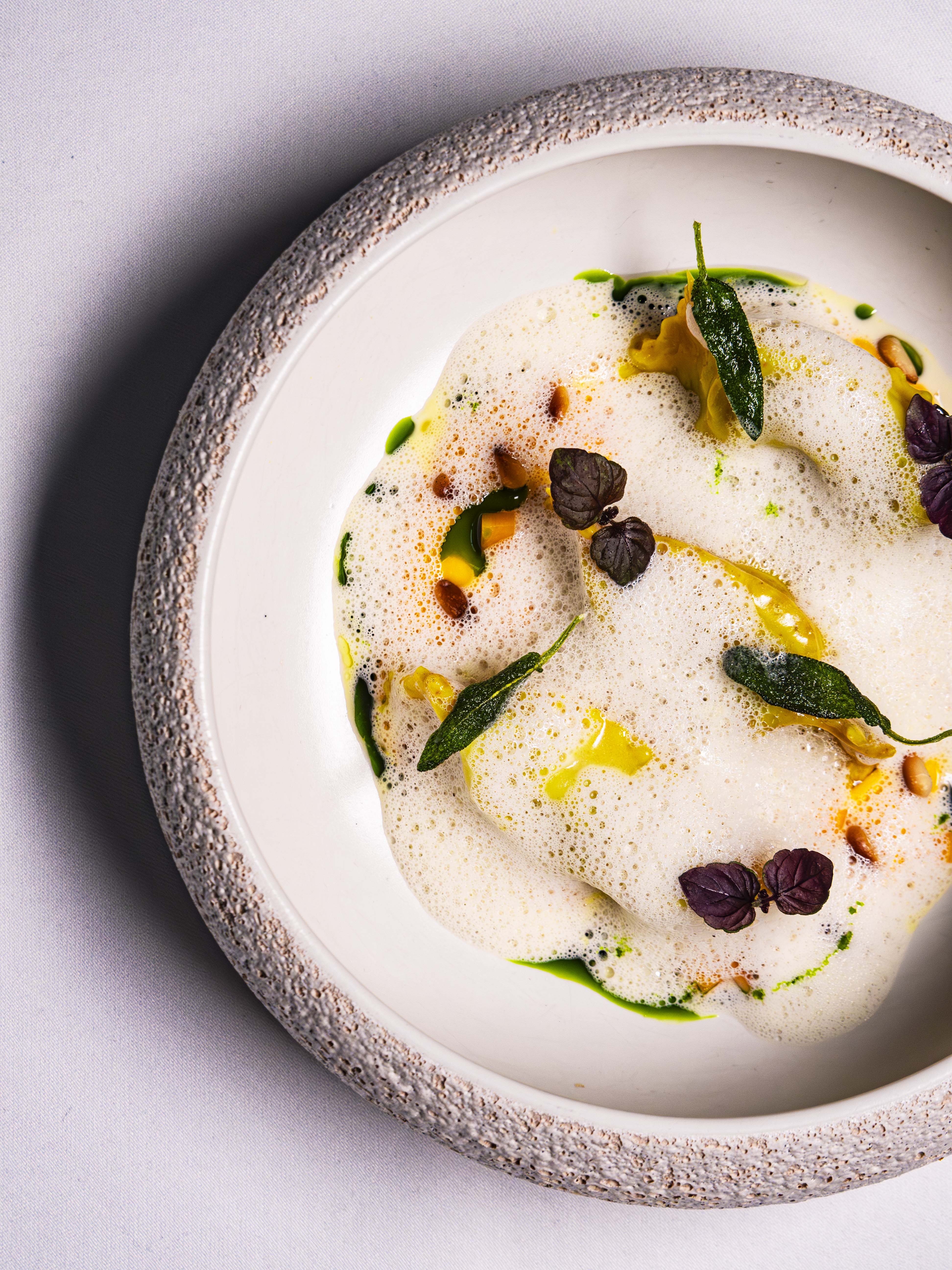 Le Cordon Bleu London - A close-up shot of a dish topped with foam, pine nuts, and fresh herbs like sage and basil. The dish is presented in a simple, elegant white bowl with a slightly textured edge.