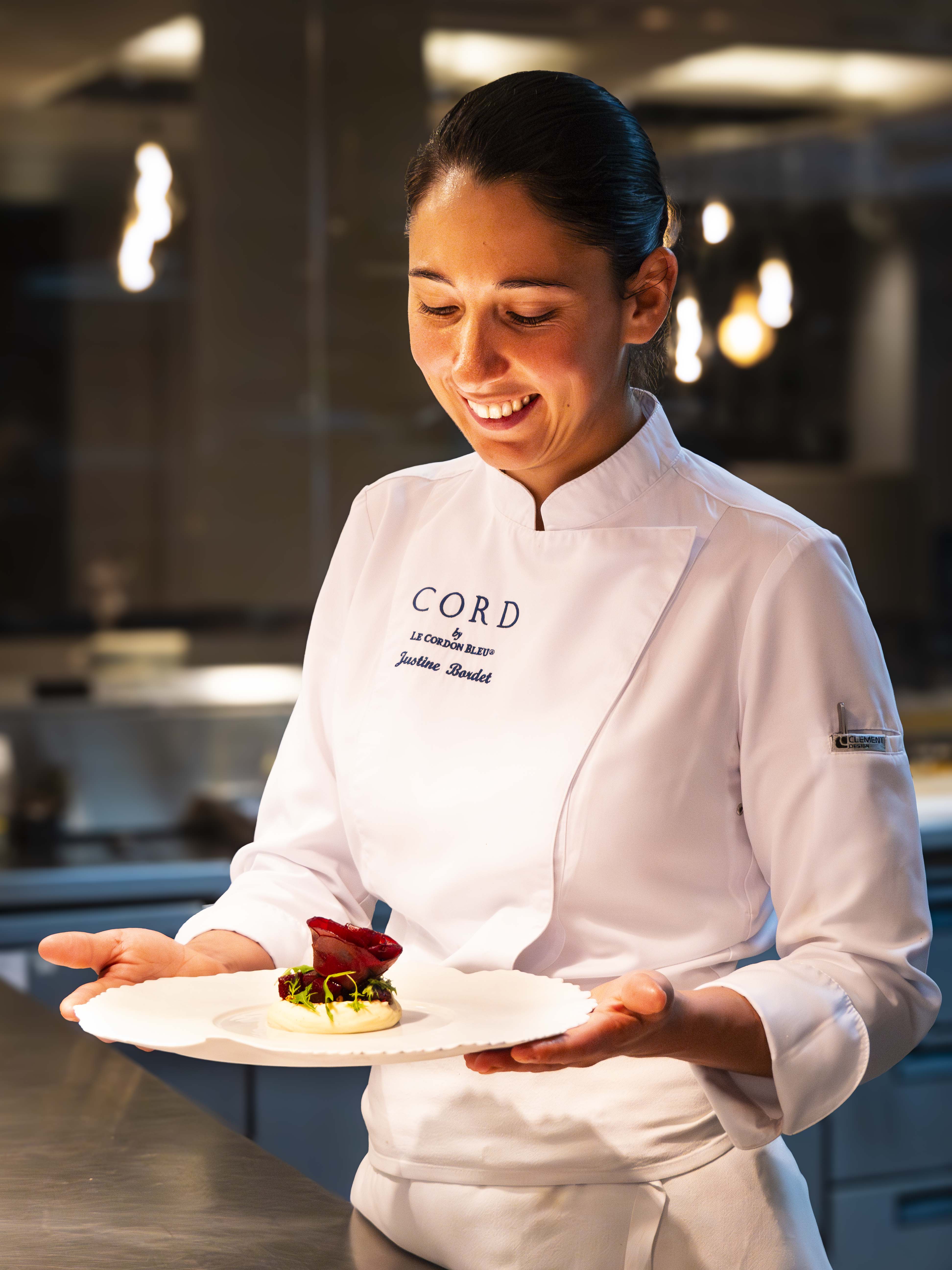 Le Cordon Bleu London - A chef, wearing a white jacket with "CORD" and "Justine Bordet" embroidered on it, is smiling while presenting a beautifully plated dish. The dish features a small portion of food, topped with a vibrant red garnish, set on a delicate white plate. The background shows a professional kitchen with soft lighting, emphasising the chef's joyful expression as she admires her creation.