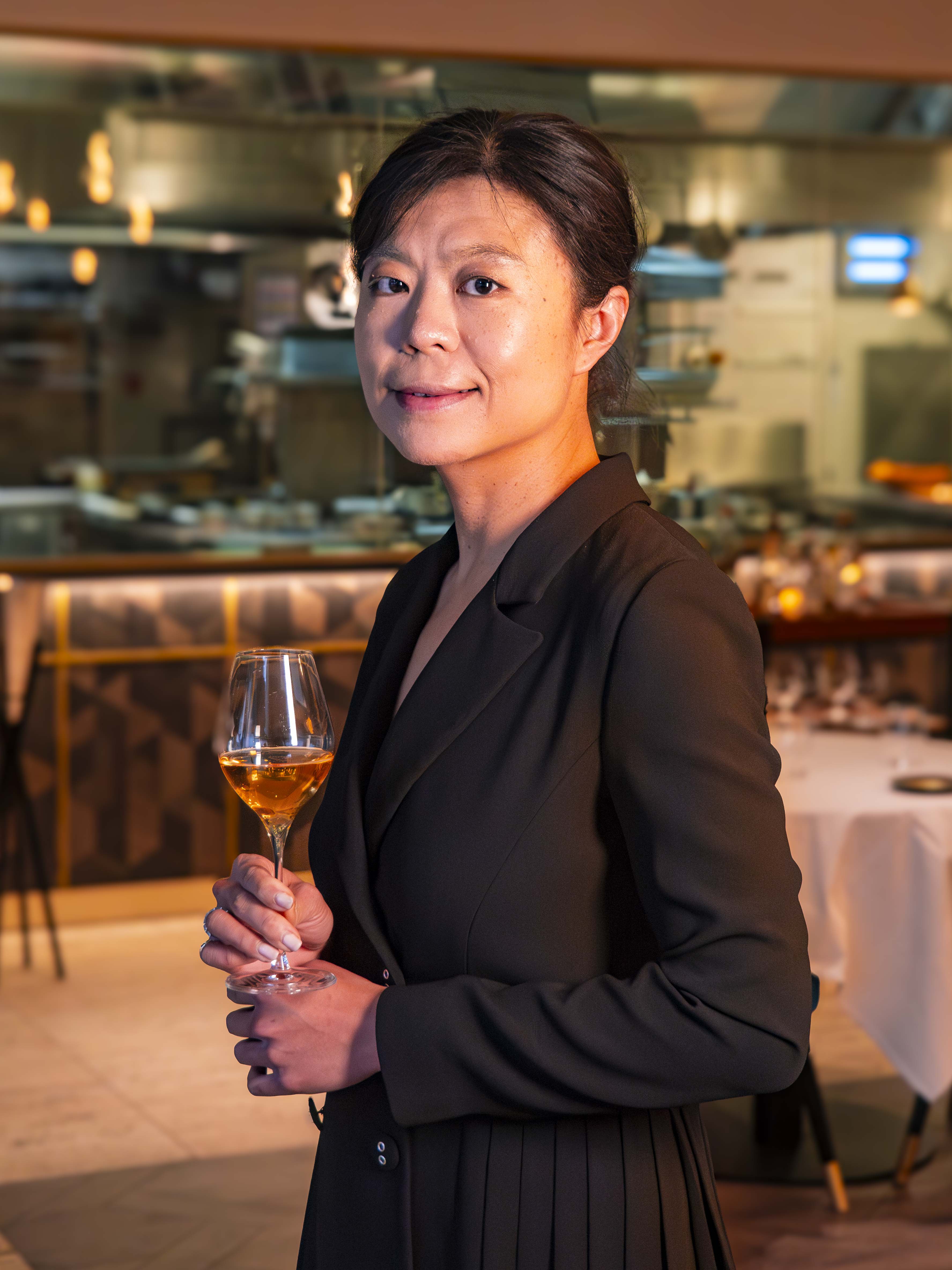 Le Cordon Bleu London - Jiachen Lu in a black blazer is holding a glass of wine and smelling it, with a thoughtful expression on her face. The background shows a softly lit restaurant kitchen, enhancing the elegant ambiance.