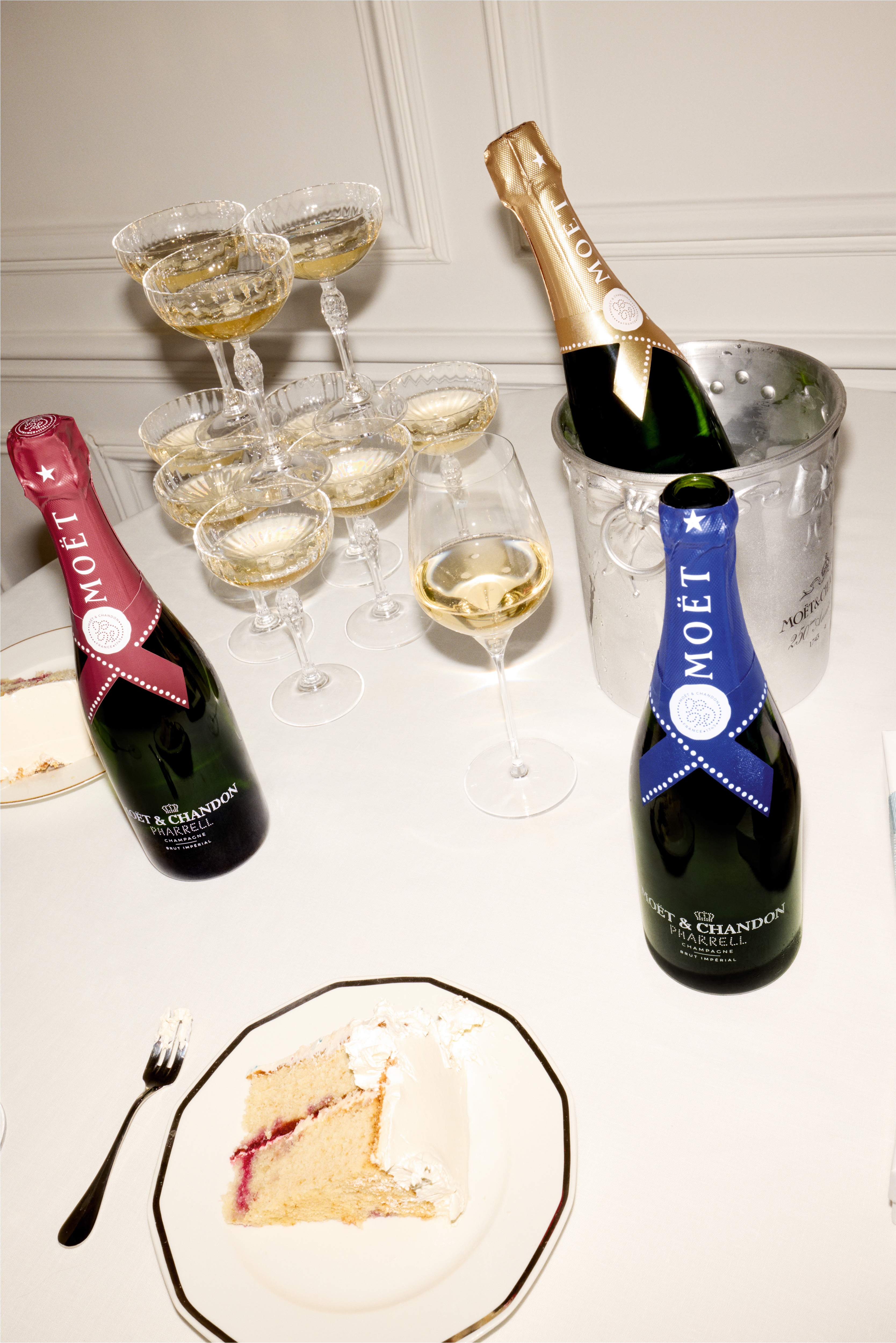 Pharrell Williams Moet Chandon - The image shows a stylish setting with two bottles of Moët & Chandon champagne, one from the Pharrell Williams limited edition collection, surrounded by elegant champagne glasses filled with bubbly. A slice of cake with a fork is also on the table, adding to the luxurious and festive atmosphere. The champagne is placed in an ice bucket, and the setting conveys an indulgent and celebratory mood.