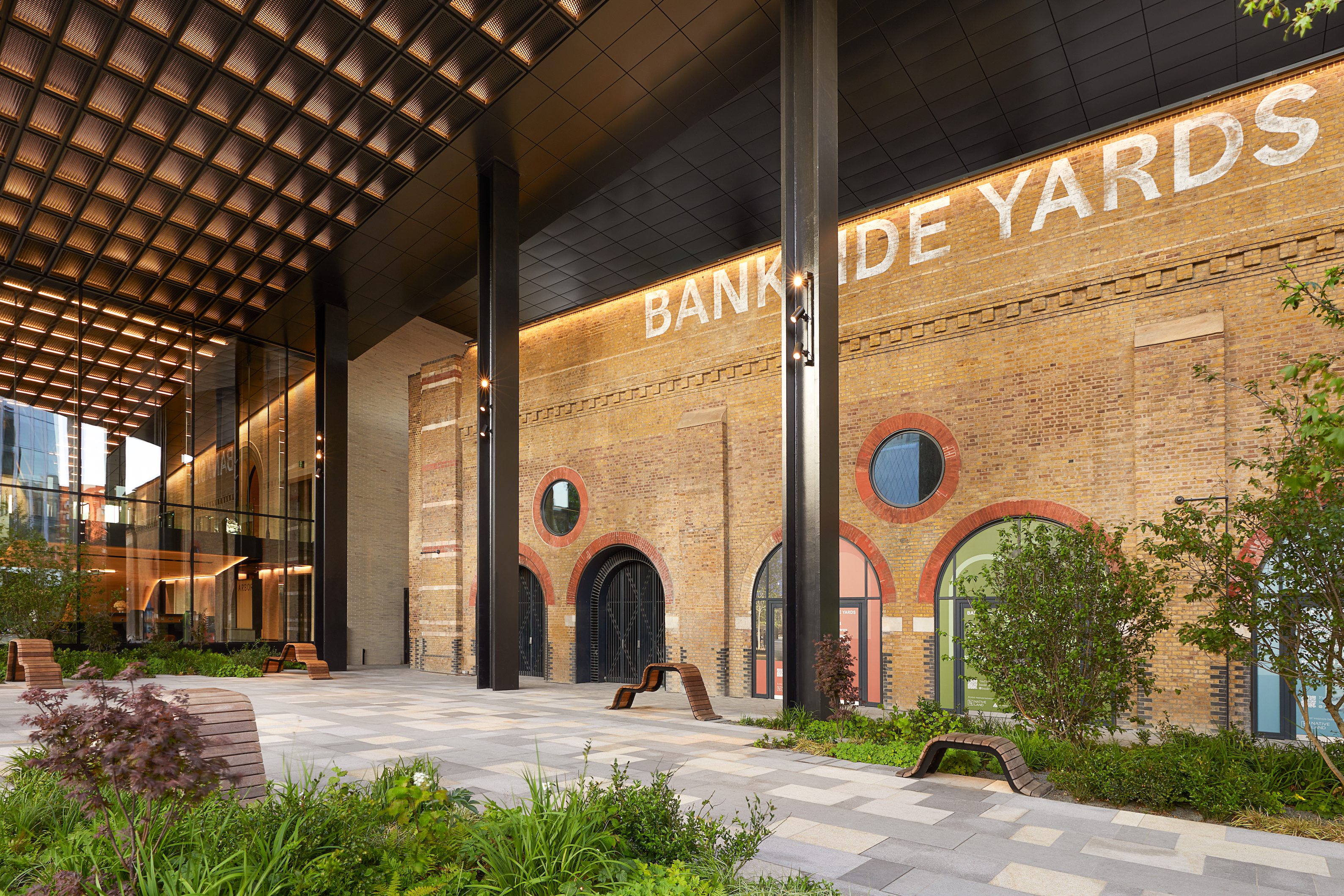 Emerging London Boroughs New Luxury Property - A modern urban courtyard with a mix of green plants and benches. The space is framed by a tall, industrial-style structure with large glass windows, and the background features a historic brick wall with round windows and the text "BANKSIDE YARDS" written in large, faded white letters.