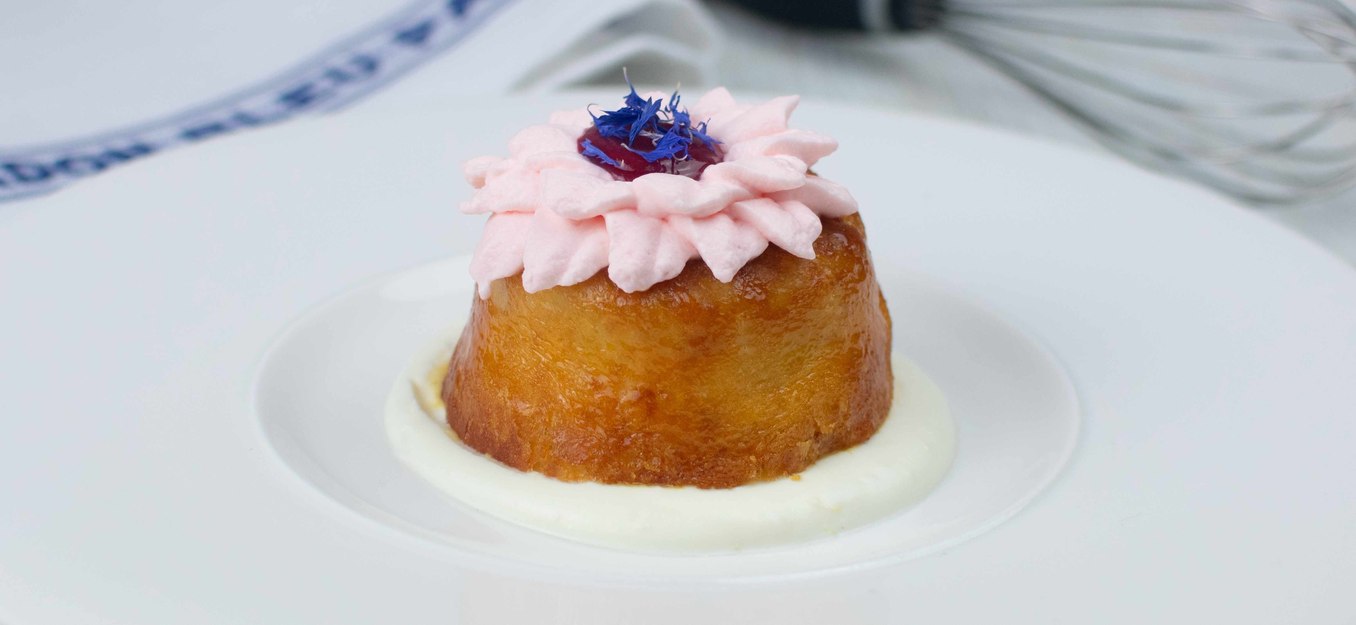 Le Cordon Bleu London - A beautifully presented dessert consisting of a small, golden cake topped with pink whipped cream, a red fruit, and purple edible flowers. The dessert is placed on a clean white plate with a creamy sauce at the base.