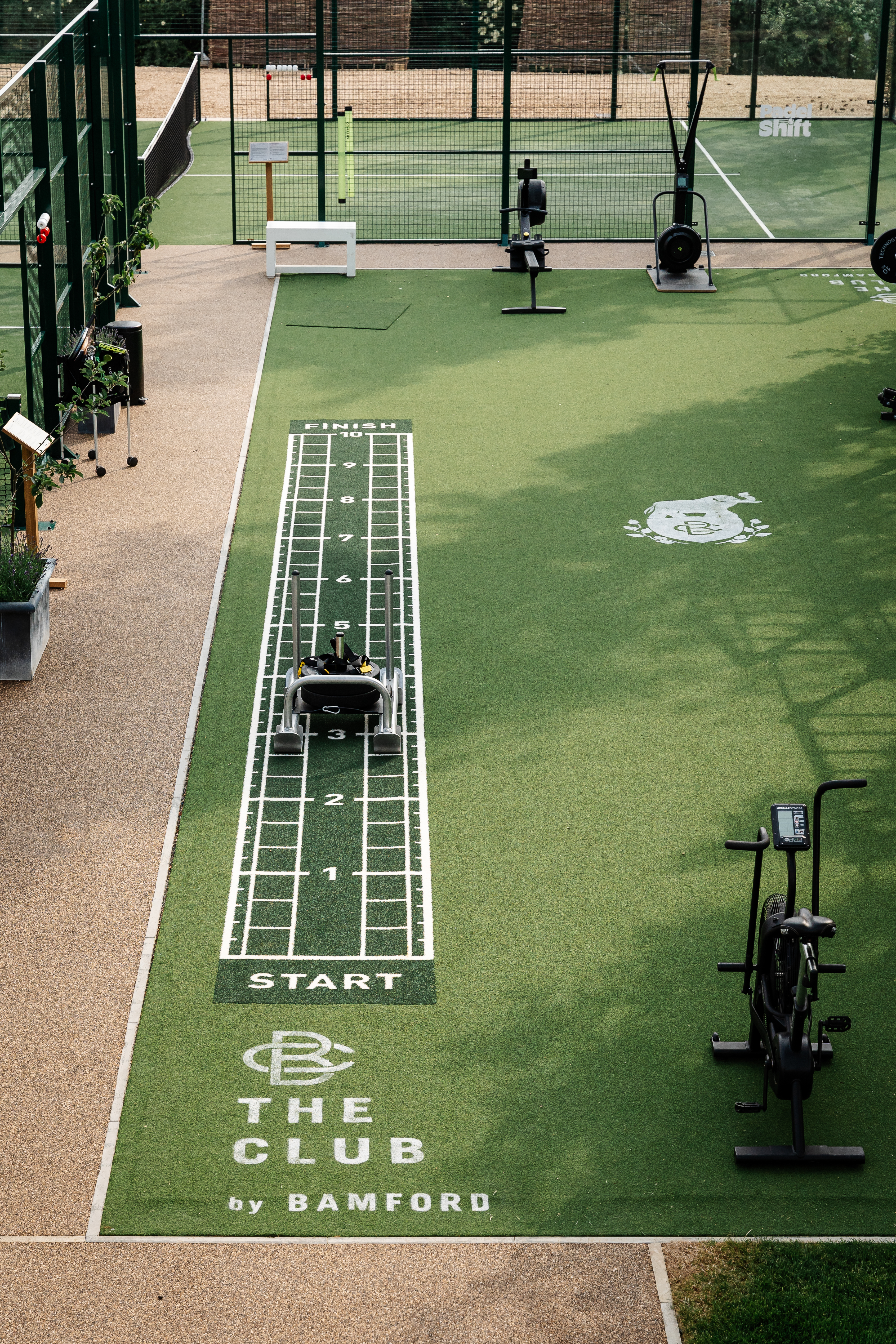 Investment, Tech & Motoring trends 2025 - An outdoor fitness area with a sled-push track, exercise equipment, and "The Club by Bamford" branding, adjacent to a tennis court