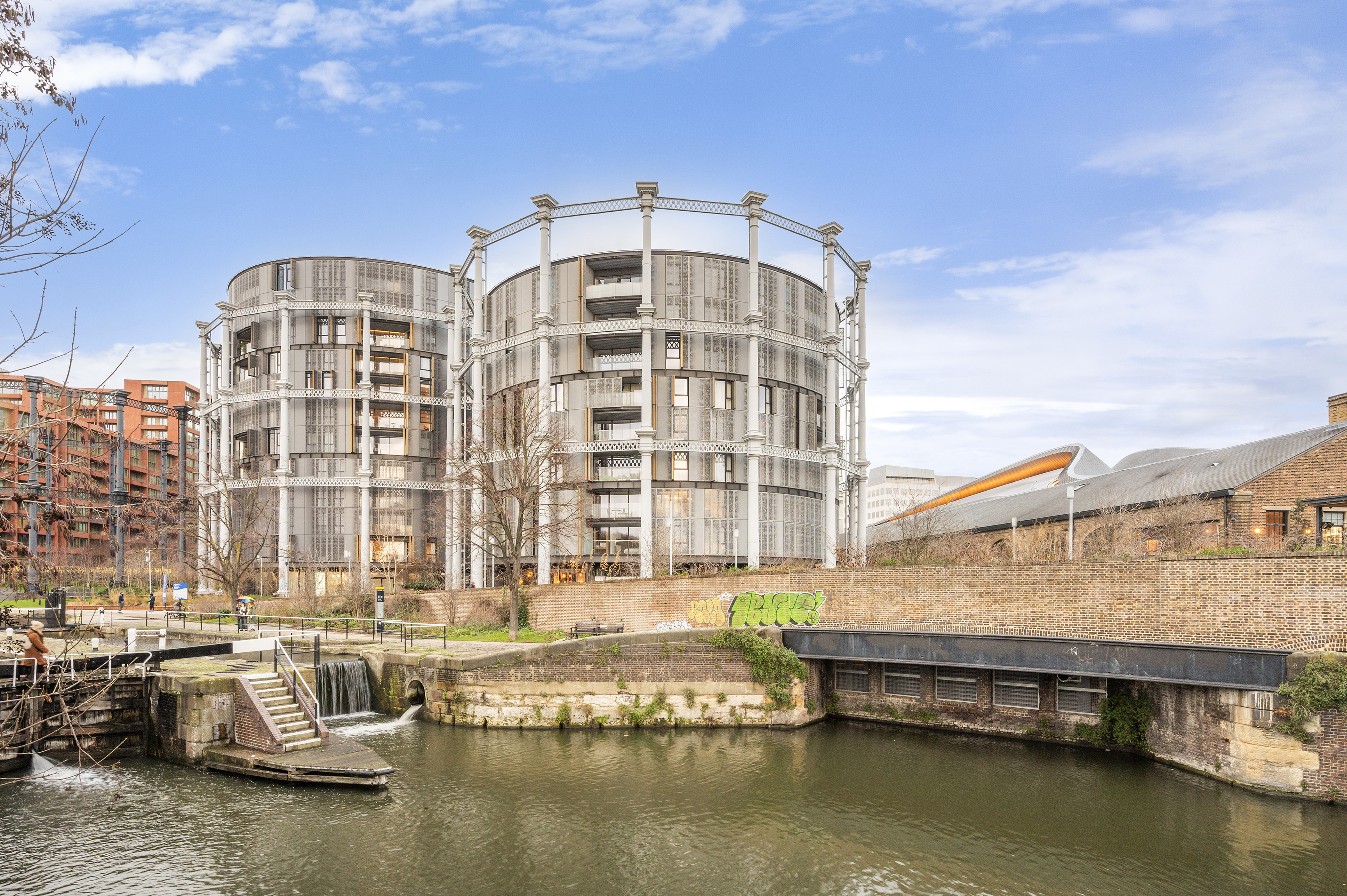 Emerging London Boroughs New Luxury Property - A modern residential building with a striking architectural design, featuring two circular structures made of metal and glass. The building overlooks a canal with green grass and a small waterfall near a staircase, while a clear sky is visible in the background. Graffiti is visible on the brick wall near the canal.