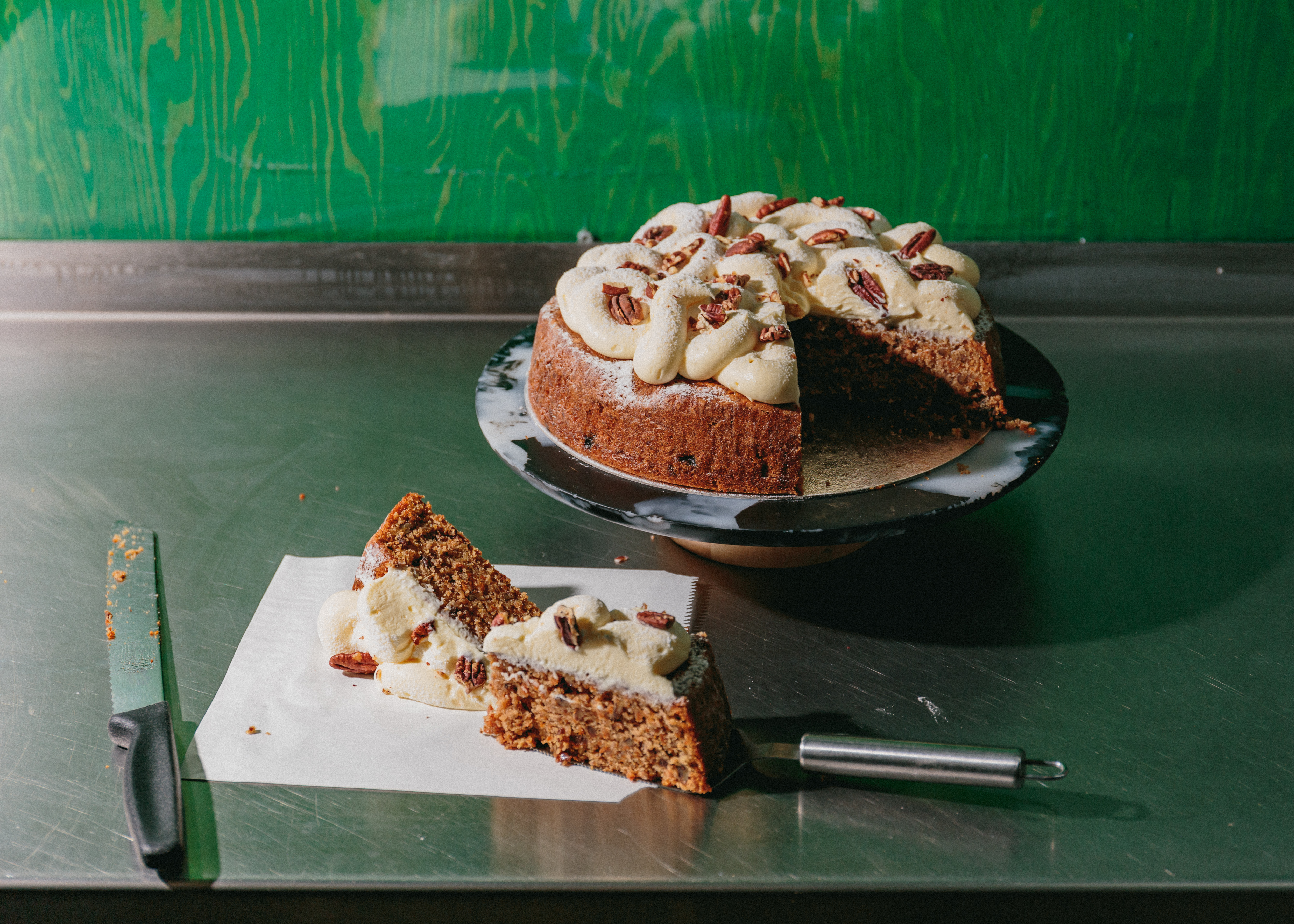 best london artisanal bakeries - A rich, moist carrot cake topped with swirls of creamy frosting and garnished with pecans is displayed on a marble cake stand. A single slice has been cut and placed on parchment paper beside the cake, revealing its texture and layers. A knife rests nearby on the stainless steel counter against a vibrant green wooden backdrop at TOAD bakery