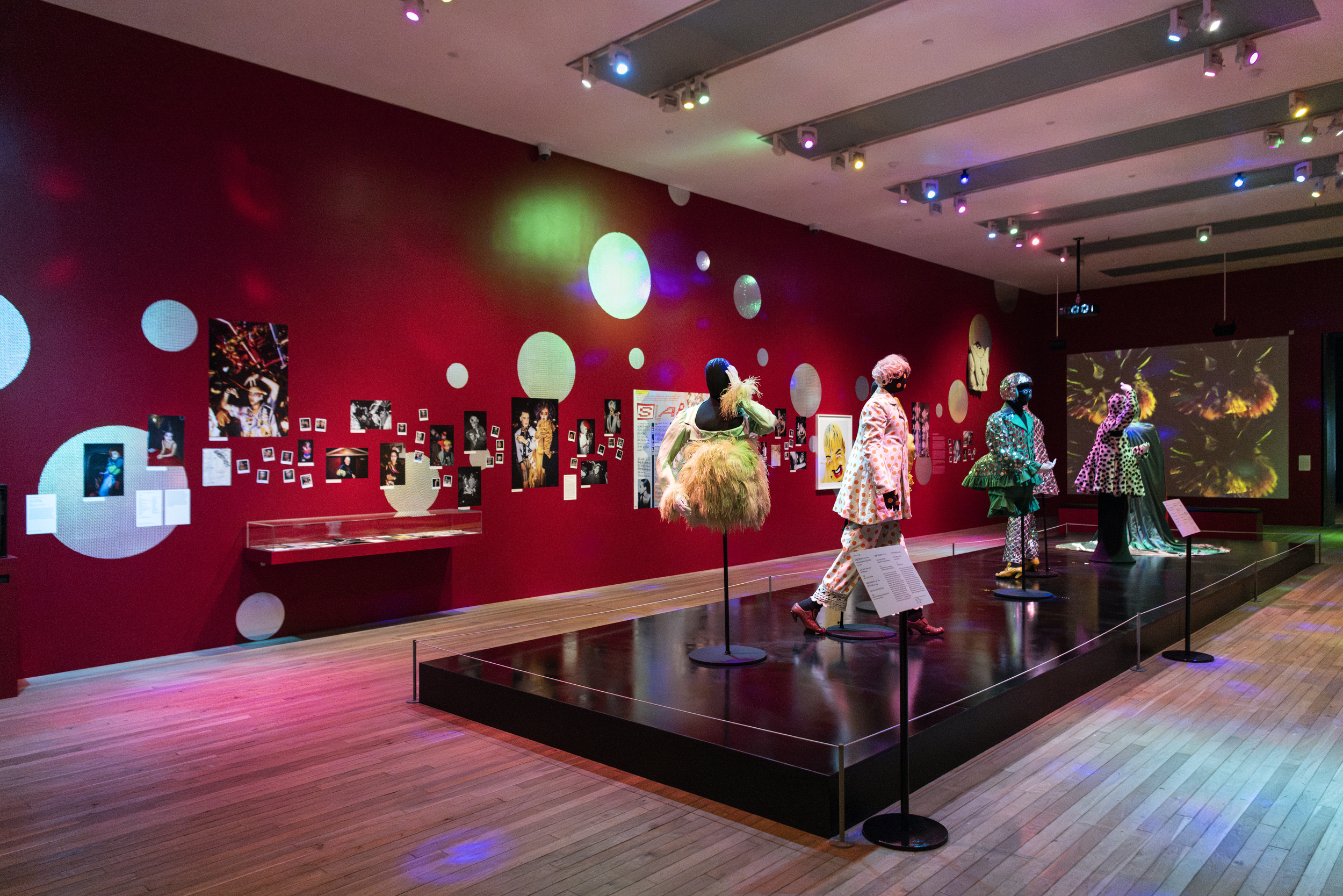 An art exhibition showcasing bold, theatrical fashion designs and photographs on a red wall. Mannequins dressed in eccentric costumes are displayed in the centre of the room, surrounded by vintage photographs and colourful lighting that creates a vibrant and immersive atmosphere