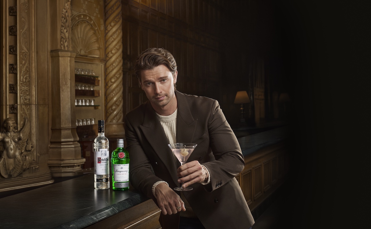 White Lotus London - A man, dressed in a brown jacket over a cream sweater, holds a martini glass in one hand while standing at a bar. Bottles of Tanqueray Gin and Ketel One Vodka are visible on the bar, with an elegant and vintage-style interior in the background, illuminated softly.