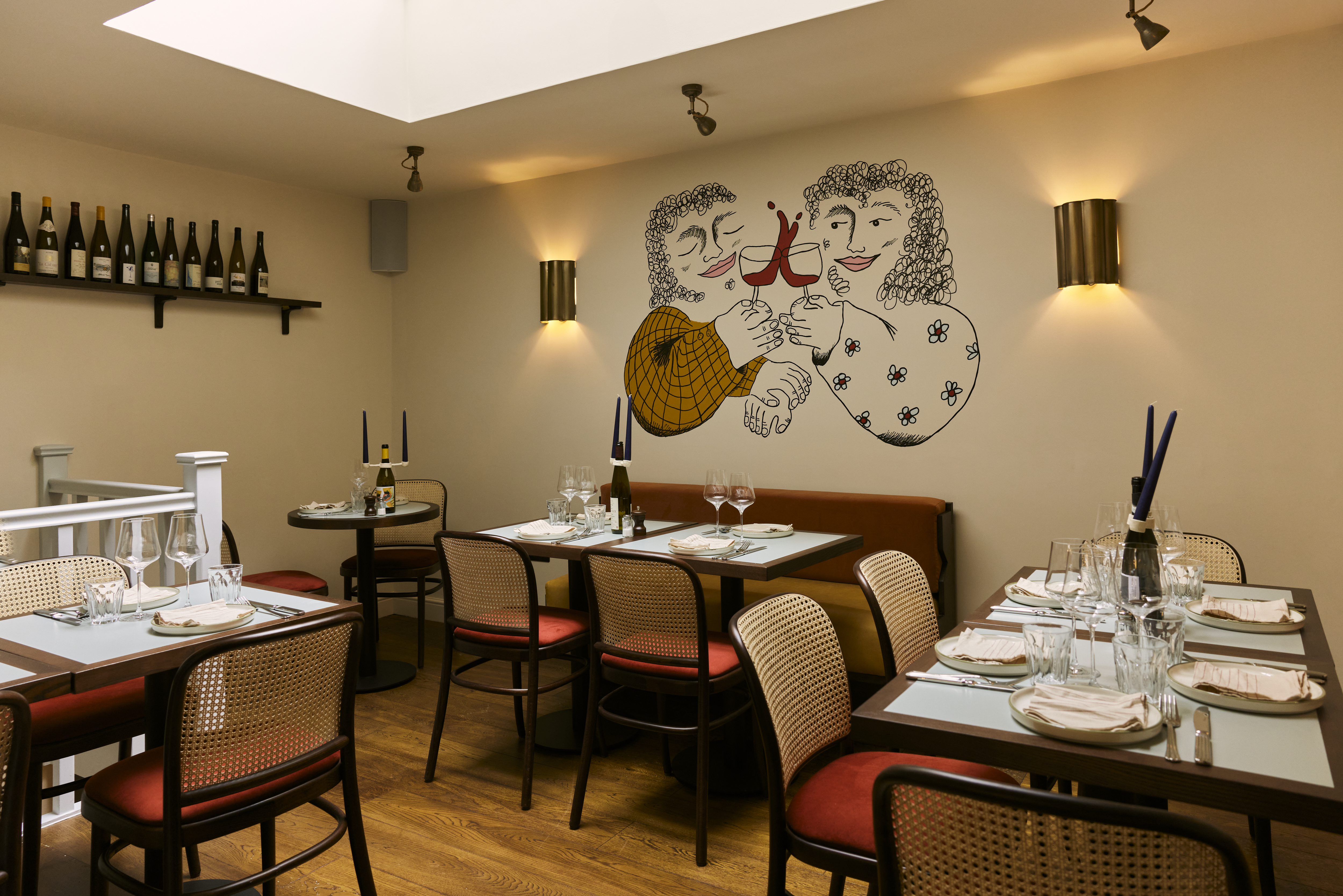 Art restaurants in London - The dining area of July featuring chairs and dark wooden tables set with glassware and tableware. A playful mural can be seen on the wall depicting two smiling figures clinking wine glasses.