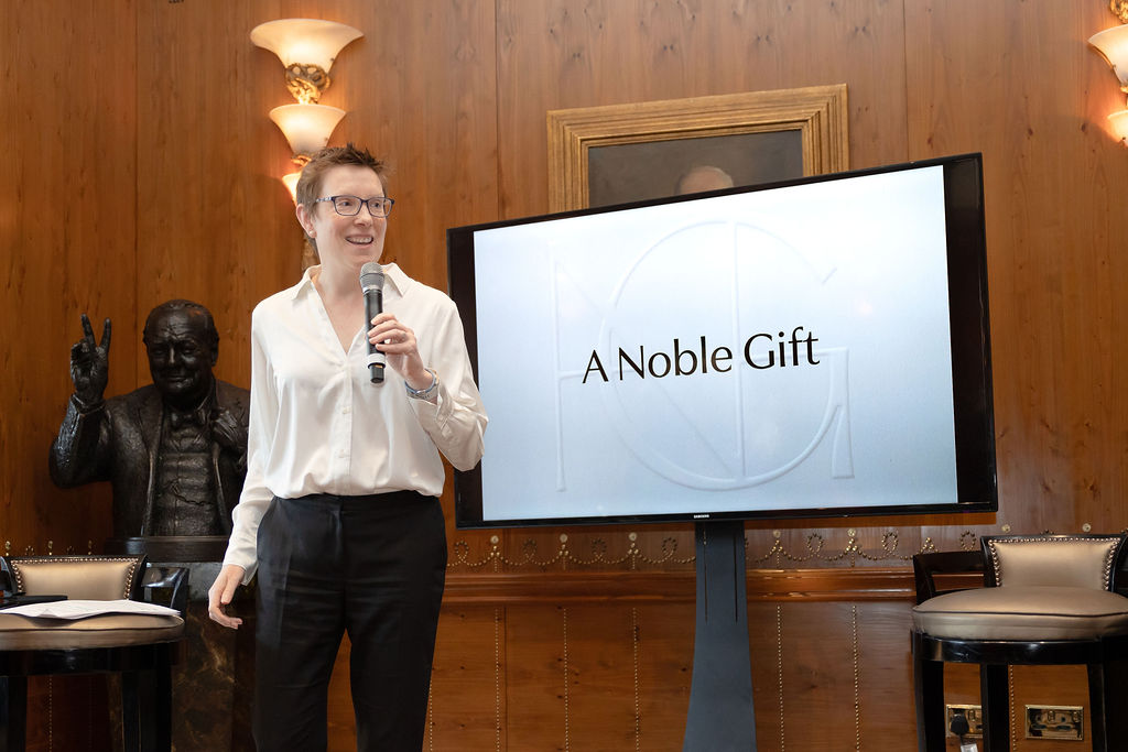 A Noble Gift Charity Auctions - Dame Tracey Crouch speaking with a microphone in front of a powerpoint presentation reading 'a noble gift'