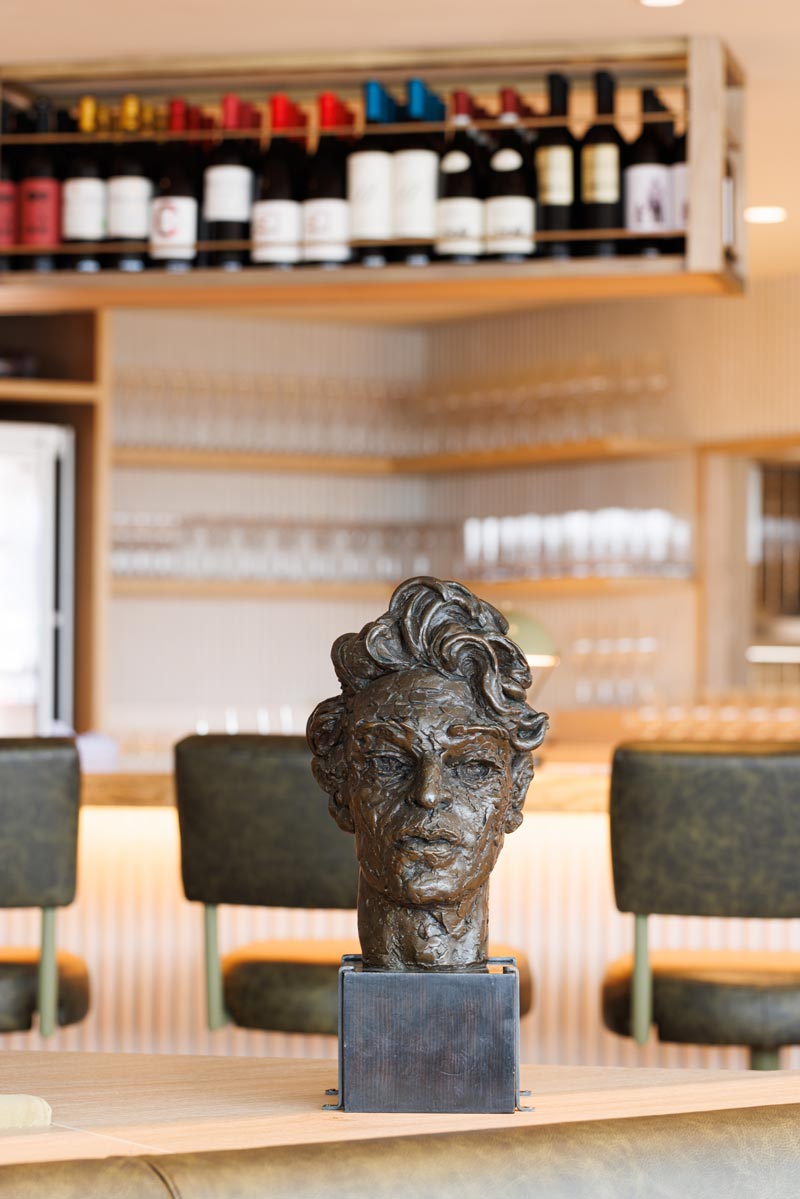 Art restaurants in London - A bronze bust sculpture of Adrian Stokes at The Portrait Restaurant displayed on a wooden surface in a bar setting. Behind this is a wine rack filled with bottles and glassware.