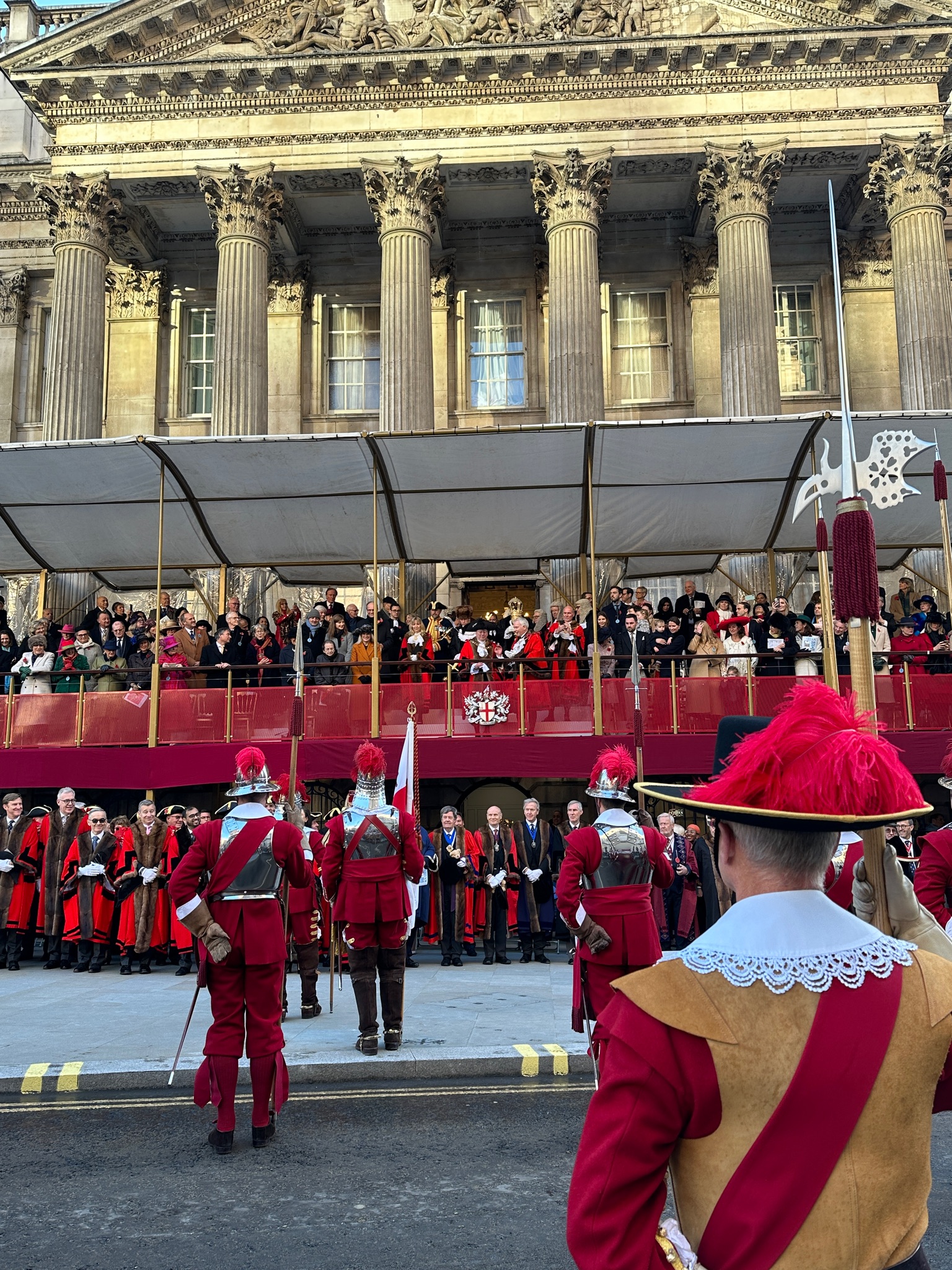 Inside Mansion House Lord Mayor Residence - The image shows a ceremonial event in front of Mansion House, with people gathered on a raised platform. In the foreground, several guards dressed in red uniforms with feathered hats stand at attention. The crowd on the balcony watches as dignitaries in formal attire are lined up. The backdrop features the grand classical architecture of Mansion House.