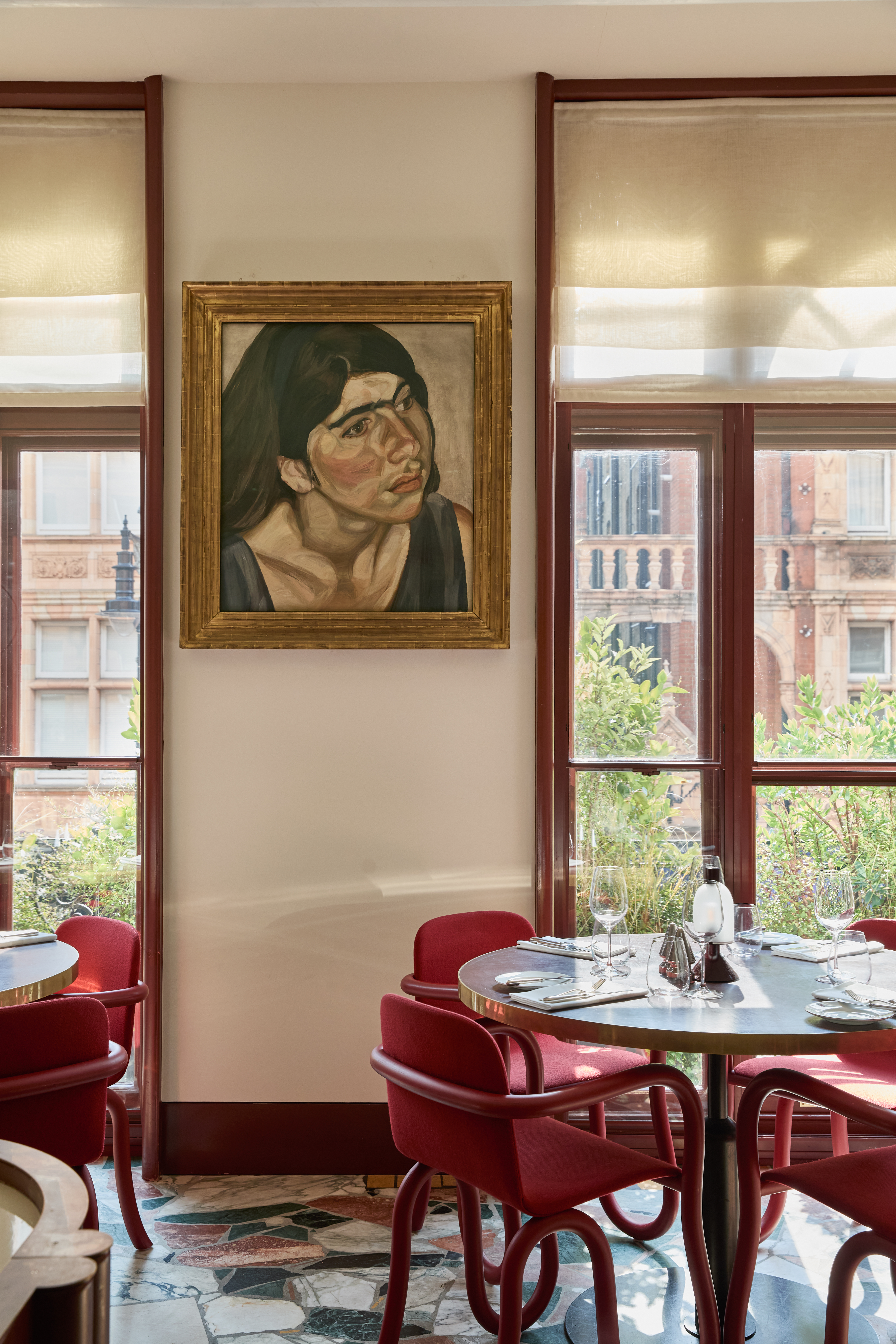 Art restaurants in London - A dining area in Mount St. Restaurant featuring a framed portrait painting of a woman by Lucian Freud. The space includes a round table set with glassware and tableware, surrounded by red chairs.