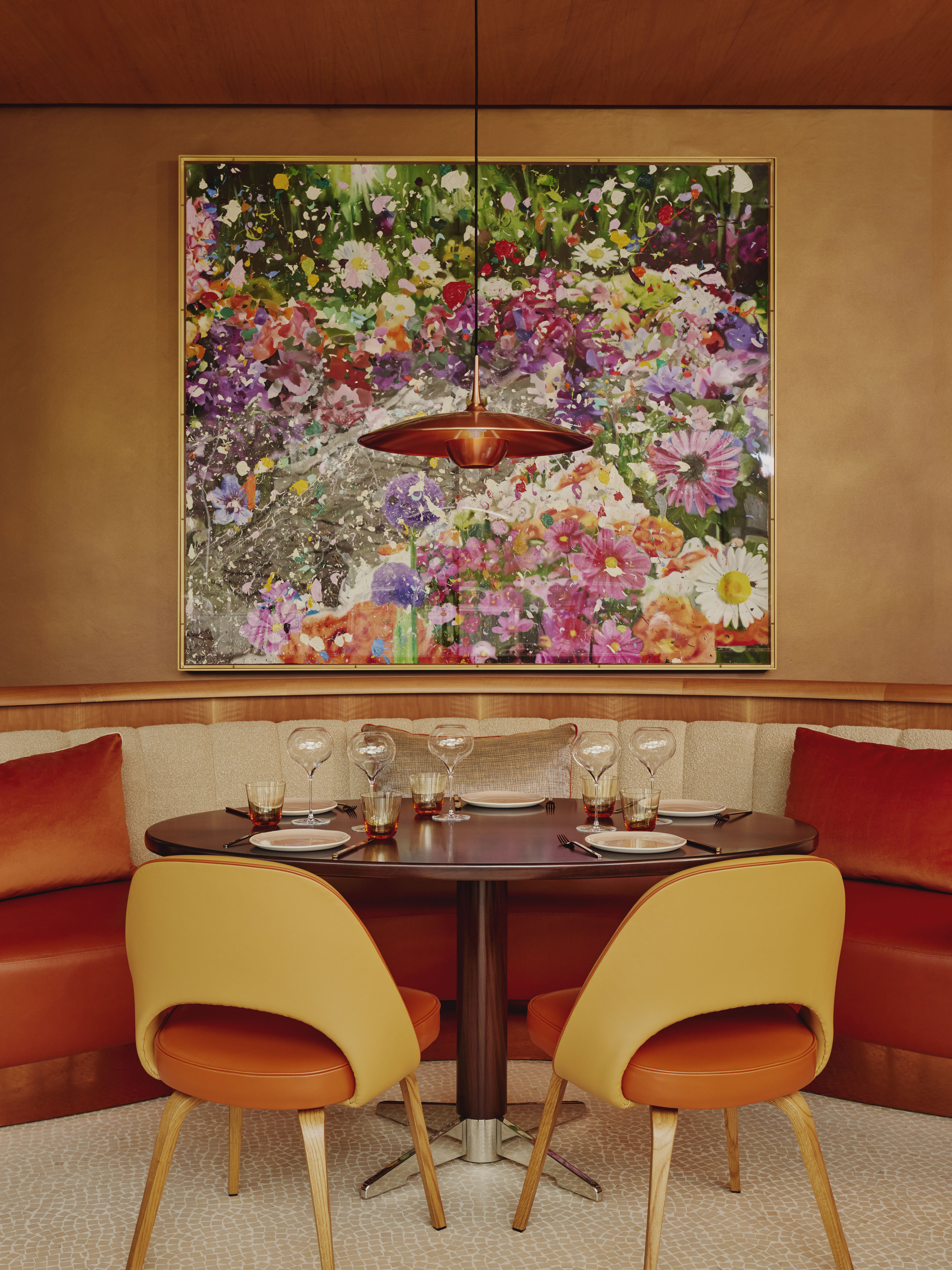 Art restaurants in London - The cosy dining area of abc kitchens at The Emory with a round wooden table, mustard-yellow chairs, and a curved banquette. The Sharing Garden (2023) by Damien Hirst hangs above.